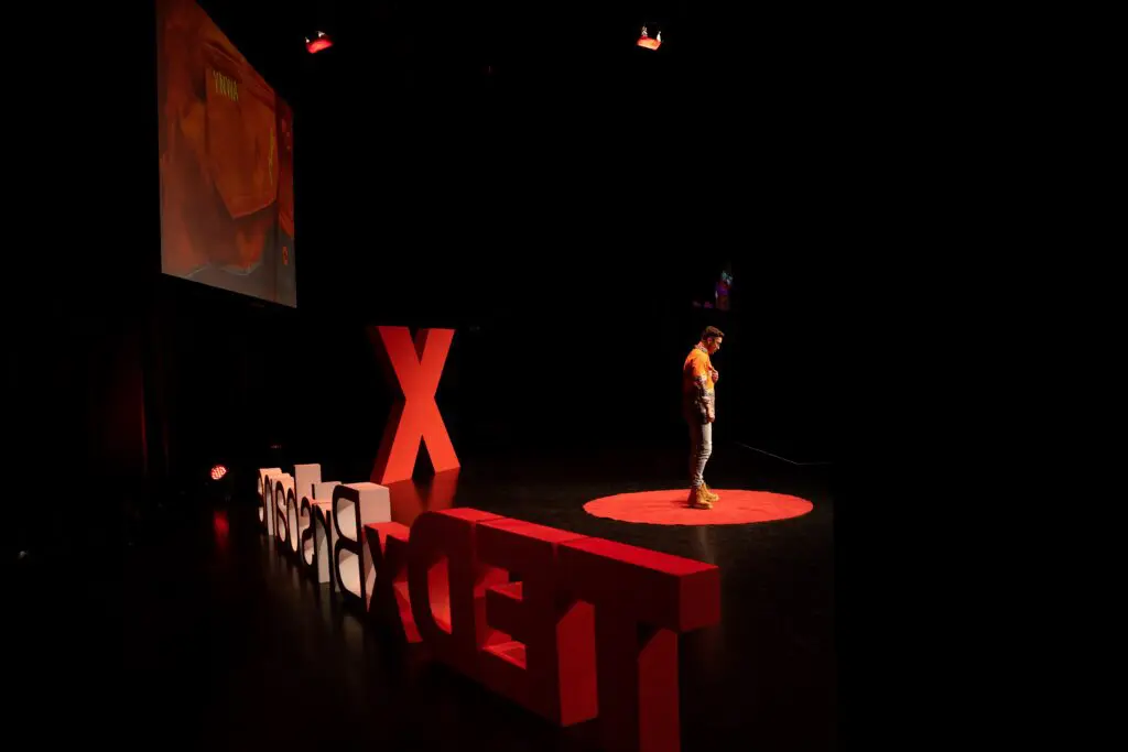 Looking from side stage wings to speaker Daniel Allen standing in the red circle on stage at TEDxBrisbane 2022 in a pensive moment with his hand pointing to his chest.