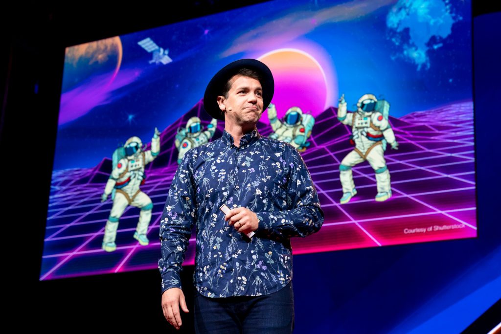 Anthony Veneziale speaks at TEDSummit: A Community Beyond Borders. July 21-25, 2019, Edinburgh, Scotland. Anthony stands in front of a slide featuring four cartoon astronauts dancing on a Tron-style planet surface.