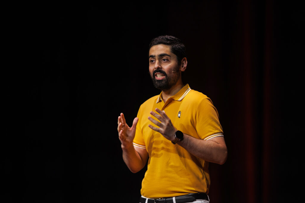 Danish Kazmi wearing a yellow polo short delivering his TEDx Talk with both hands raised in front of him emphasising a point.
