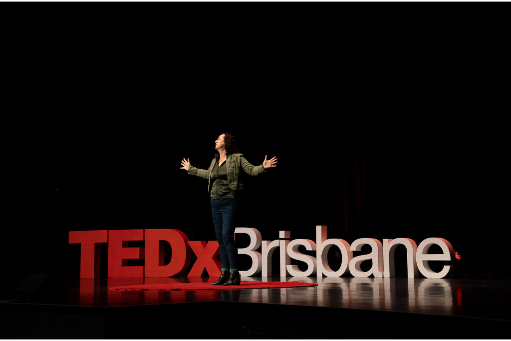 Adrienne Alexander wearing a green jacked and jeans delivering her TEDx Talk with both hands, open palms at 90 degrees to her side with palms facing front emphasising a point.